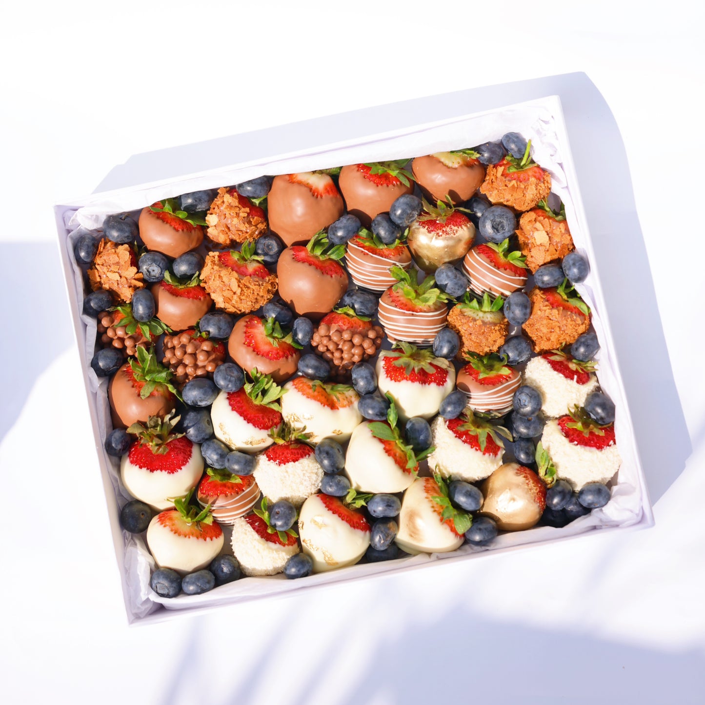 A neatly arranged dessert box featuring a variety of chocolate-covered strawberries (milk, white, and decorated), golden-dusted strawberries, crunchy caramel bites, blueberries, and small chocolate crisps, all placed in a white box with a tissue paper lining.