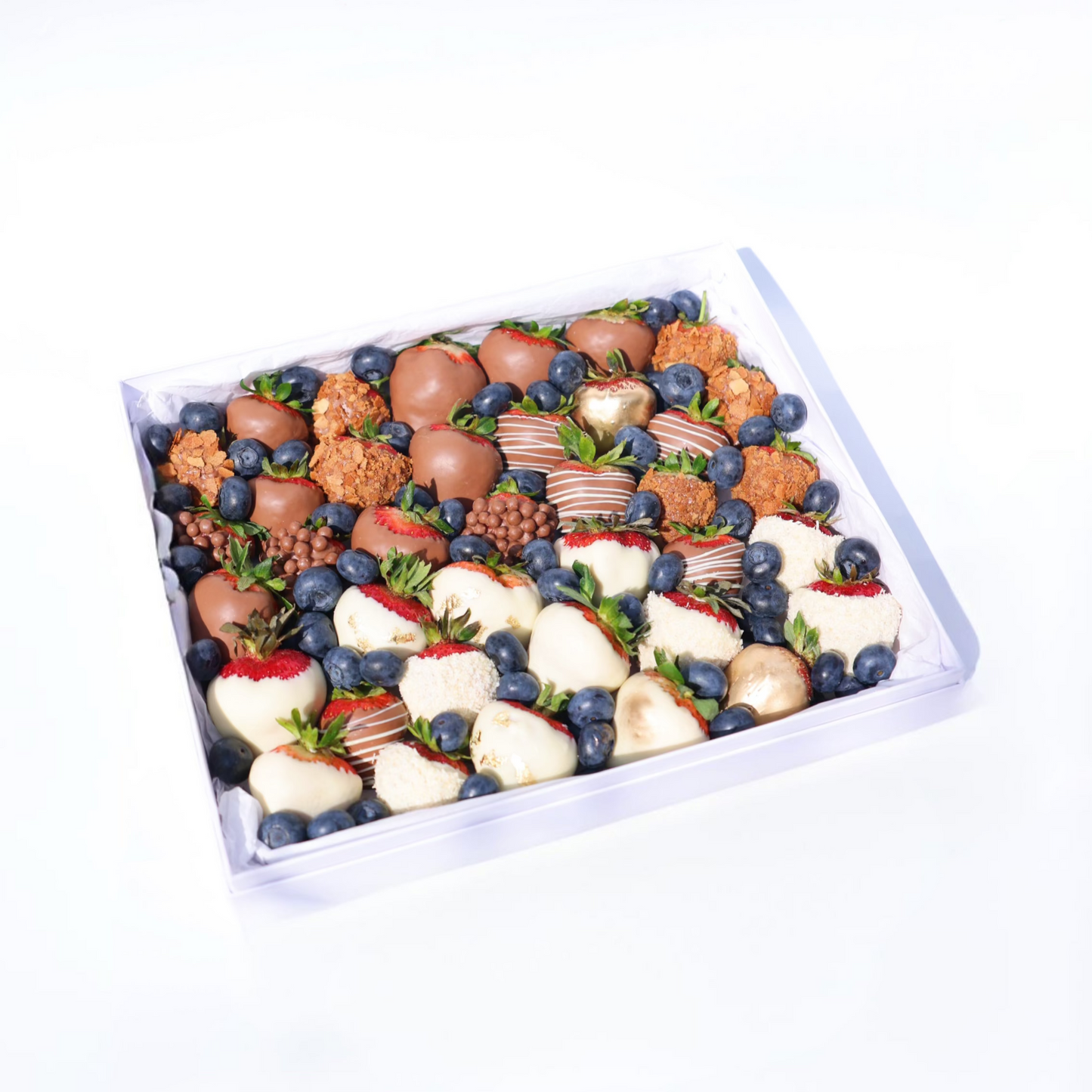 A neatly arranged dessert box featuring a variety of chocolate-covered strawberries (milk, white, and decorated), golden-dusted strawberries, crunchy caramel bites, blueberries, and small chocolate crisps, all placed in a white box with a tissue paper lining.