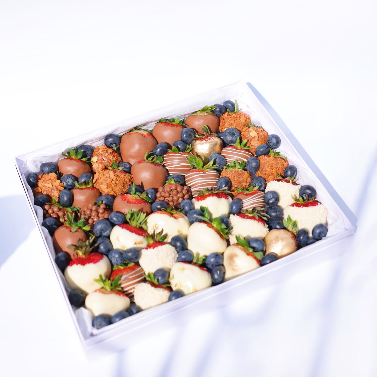 A neatly arranged dessert box featuring a variety of chocolate-covered strawberries (milk, white, and decorated), golden-dusted strawberries, crunchy caramel bites, blueberries, and small chocolate crisps, all placed in a white box with a tissue paper lining.