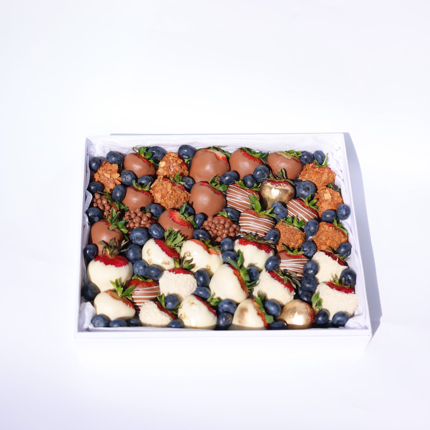 A neatly arranged dessert box featuring a variety of chocolate-covered strawberries (milk, white, and decorated), golden-dusted strawberries, crunchy caramel bites, blueberries, and small chocolate crisps, all placed in a white box with a tissue paper lining.