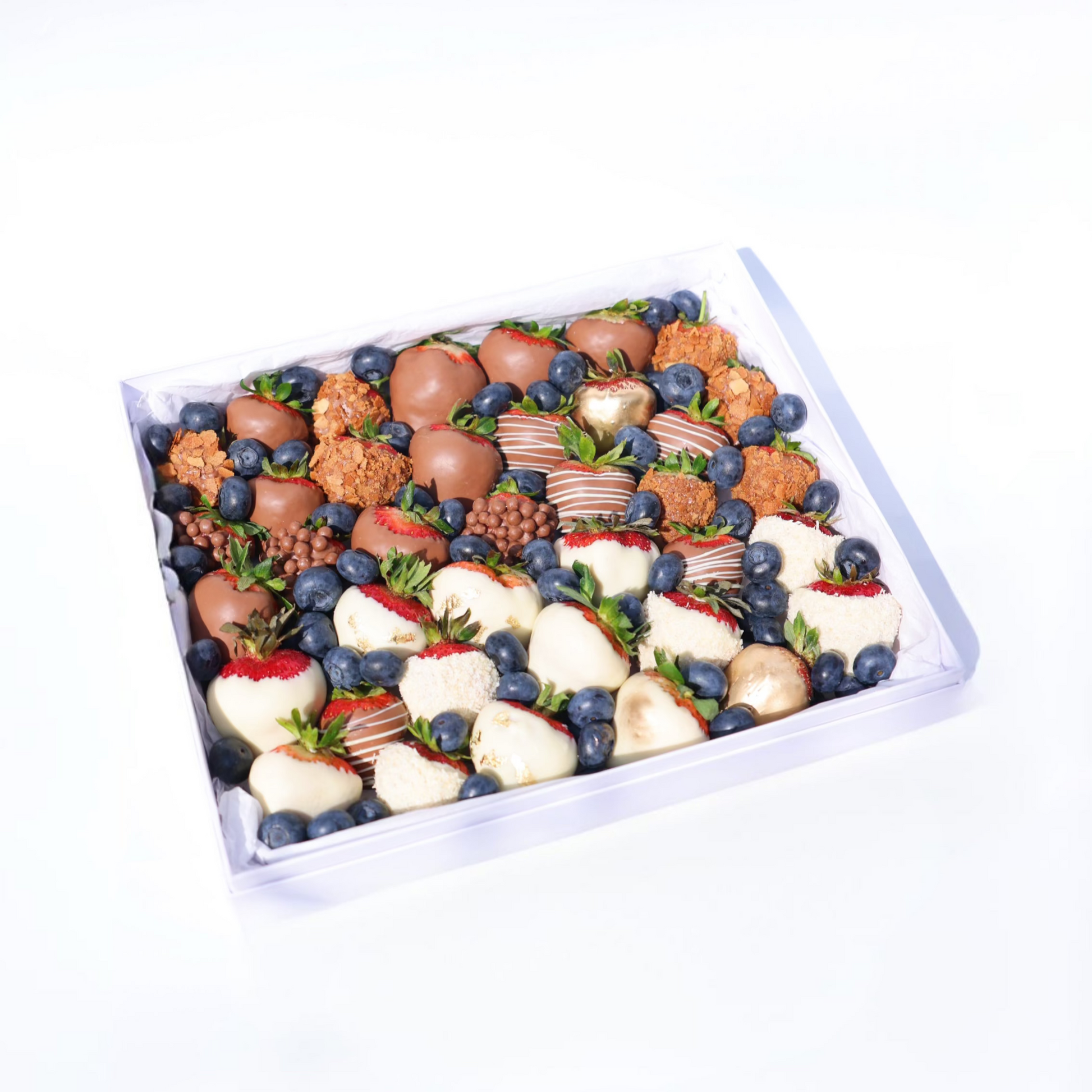 A neatly arranged dessert box featuring a variety of chocolate-covered strawberries (milk, white, and decorated), golden-dusted strawberries, crunchy caramel bites, blueberries, and small chocolate crisps, all placed in a white box with a tissue paper lining.